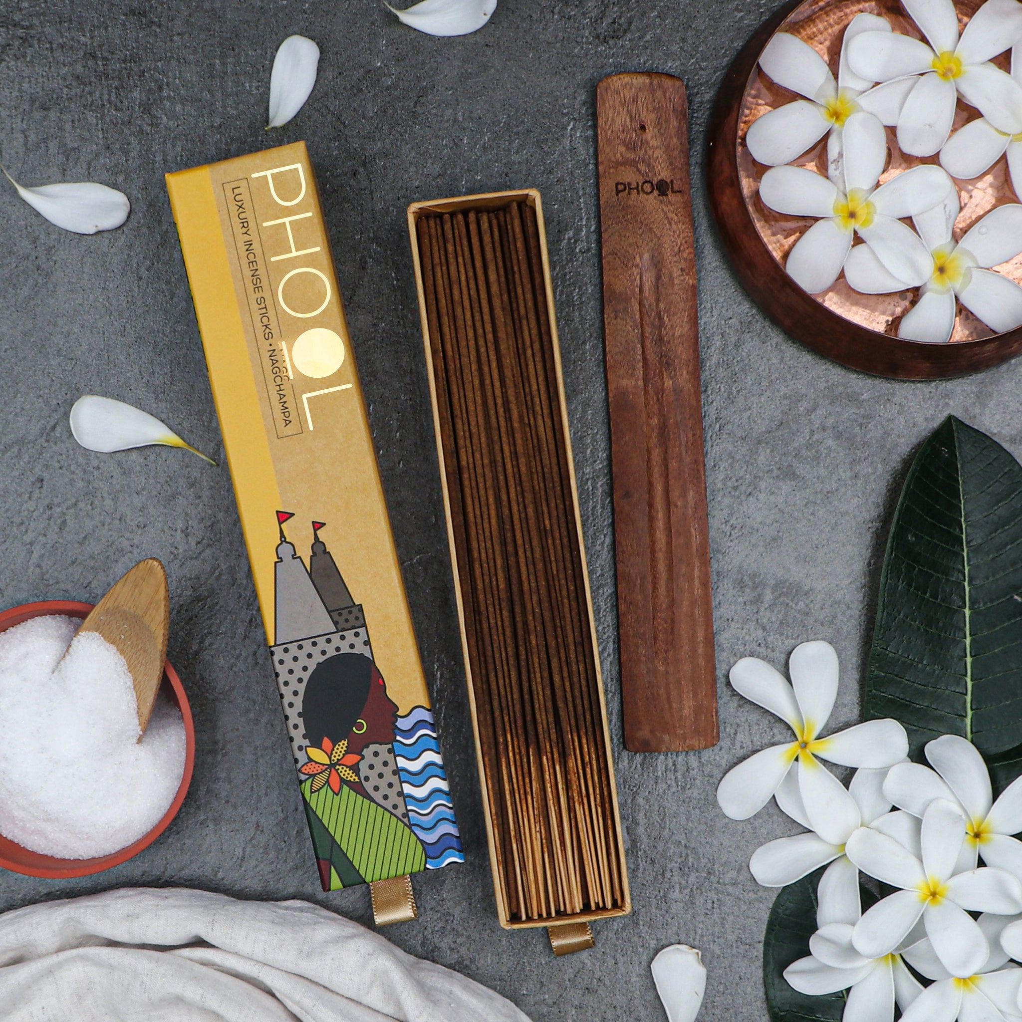 Open box of Phool Nagchampa Incense Sticks and wooden holder on grey textured surface surrounded by white flowers.