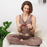 A woman sits cross-legged on a chair, meditating with Maya Shanti Yoga Tingsha cymbals, a plant and pillow nearby.