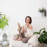 Woman sitting with legs up on cream armchair smiling towards someone off-camera holding MSY Hammered Copper Water Bottle and lid.