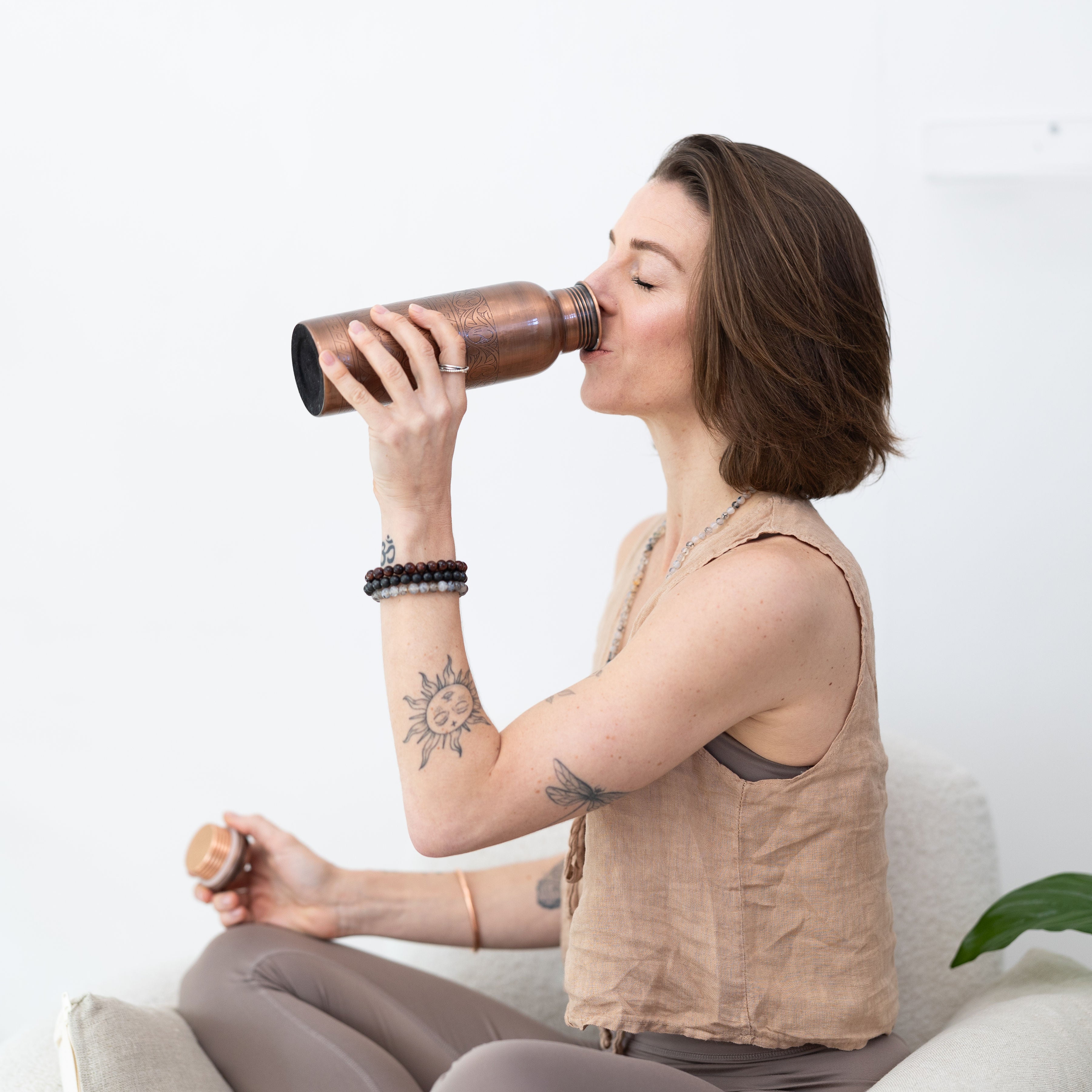 Woman sitting cross-legged drinking from a Maya Shanti Yoga Etched 750ml Copper Water Bottle with the lid in her other hand.