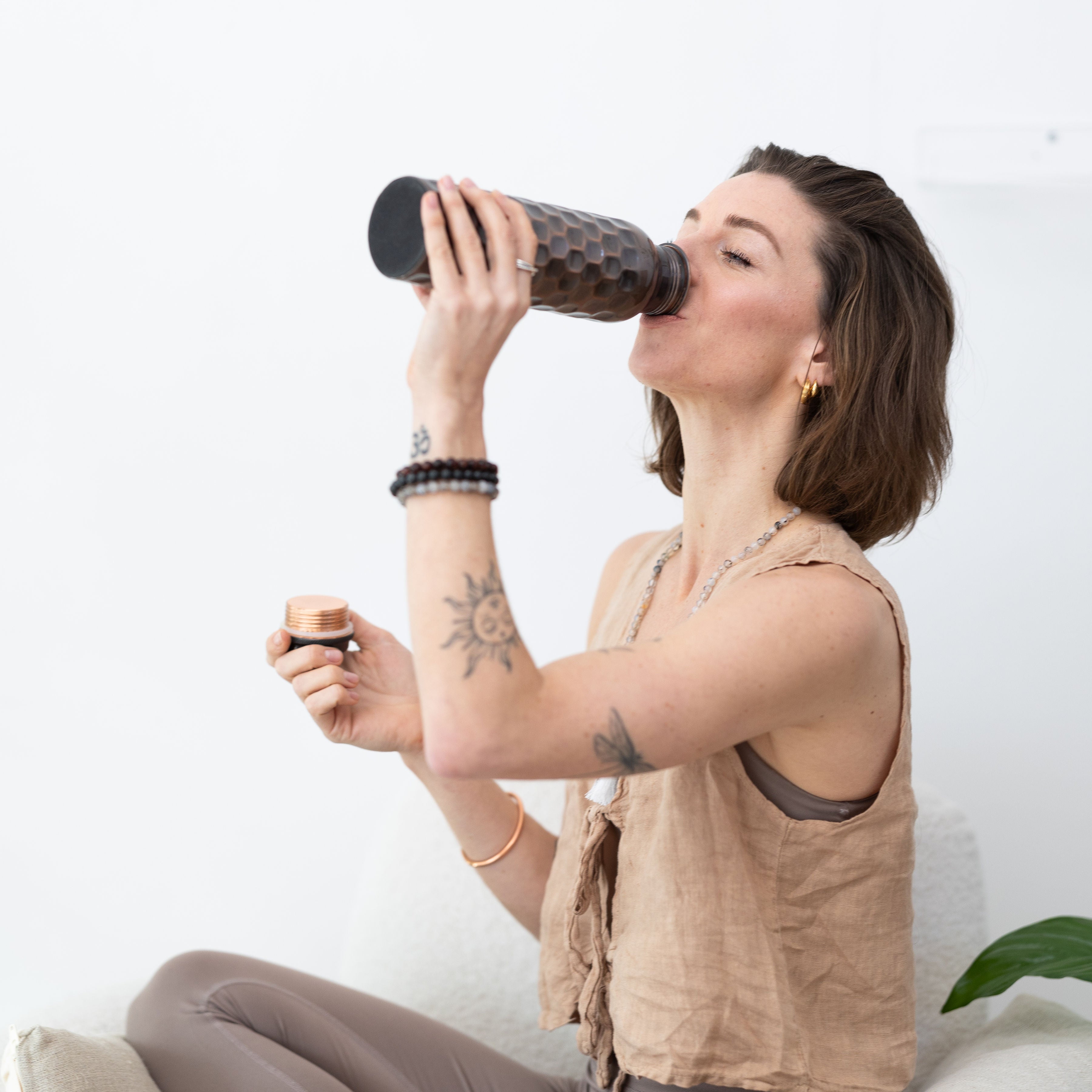 A woman sits cross-legged, drinking from a Maya Shanti Yoga Brown Antique 750ml Copper Water Bottle, holding the lid in her other hand.