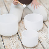 Kneeling on the wooden floor, a person holds a mallet over the Maya Shanti Yoga MSY Crystal Singing Bowls - Set of 3, creating gentle tones at 432 Hz that resonate through the room for sound healing.