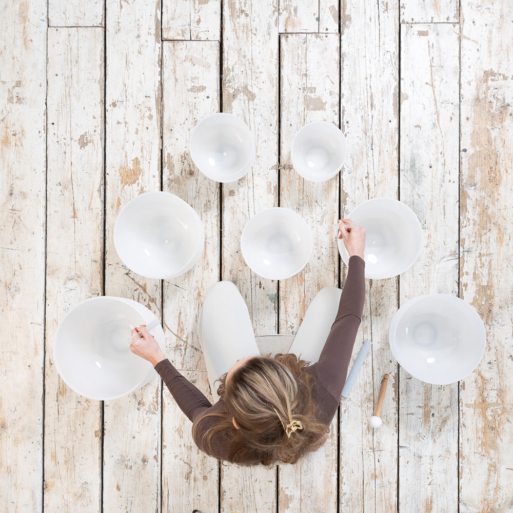 A person sits on a wooden floor using a Maya Shanti Yoga MSY Crystal Singing Bowls Set of 7 (432 Hz), playing them with mallets to create soothing vibrations for sound healing.