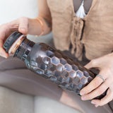 Person sits cross-legged in neutral clothing, holding a Maya Shanti Yoga Brown Antique 750ml Copper Water Bottle with a screw-on cap.
