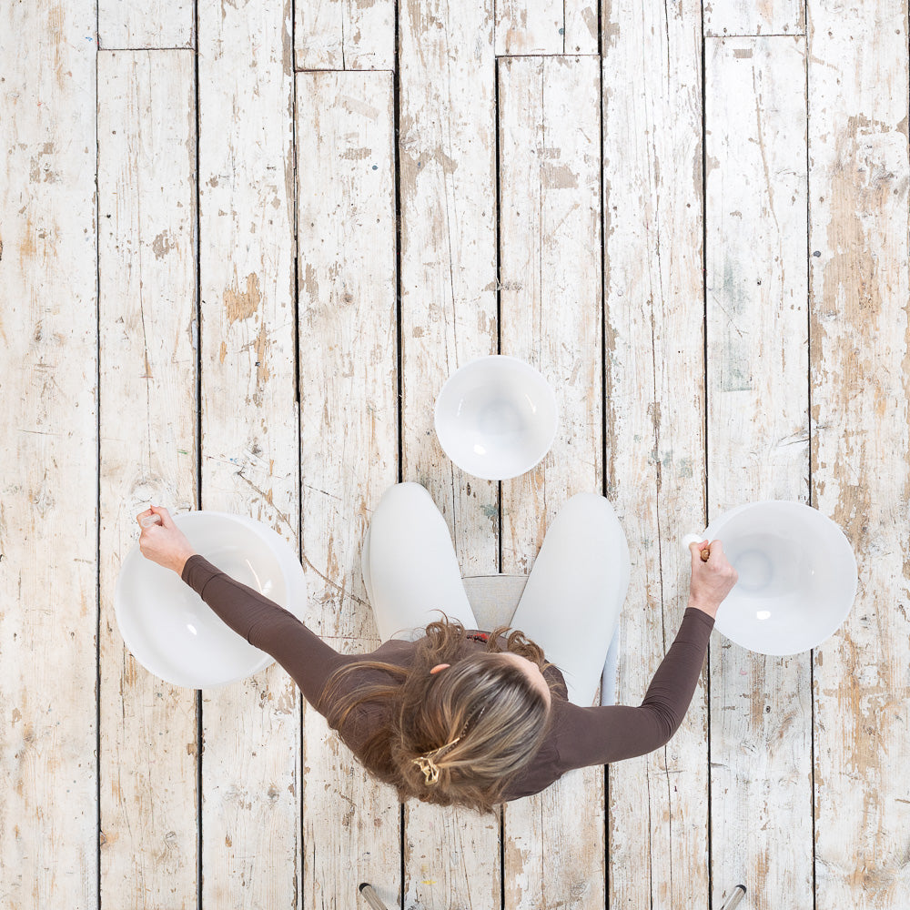 Top view of a person on a wooden floor holding two Maya Shanti Yoga MSY Crystal Singing Bowls (432 Hz), part of a 3-piece set, with the third bowl nearby, preparing for a serene sound healing session.
