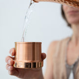 A person pours water from the Maya Shanti Yoga Hammered 950ml Copper Carafe into a pure copper cup with their left hand.