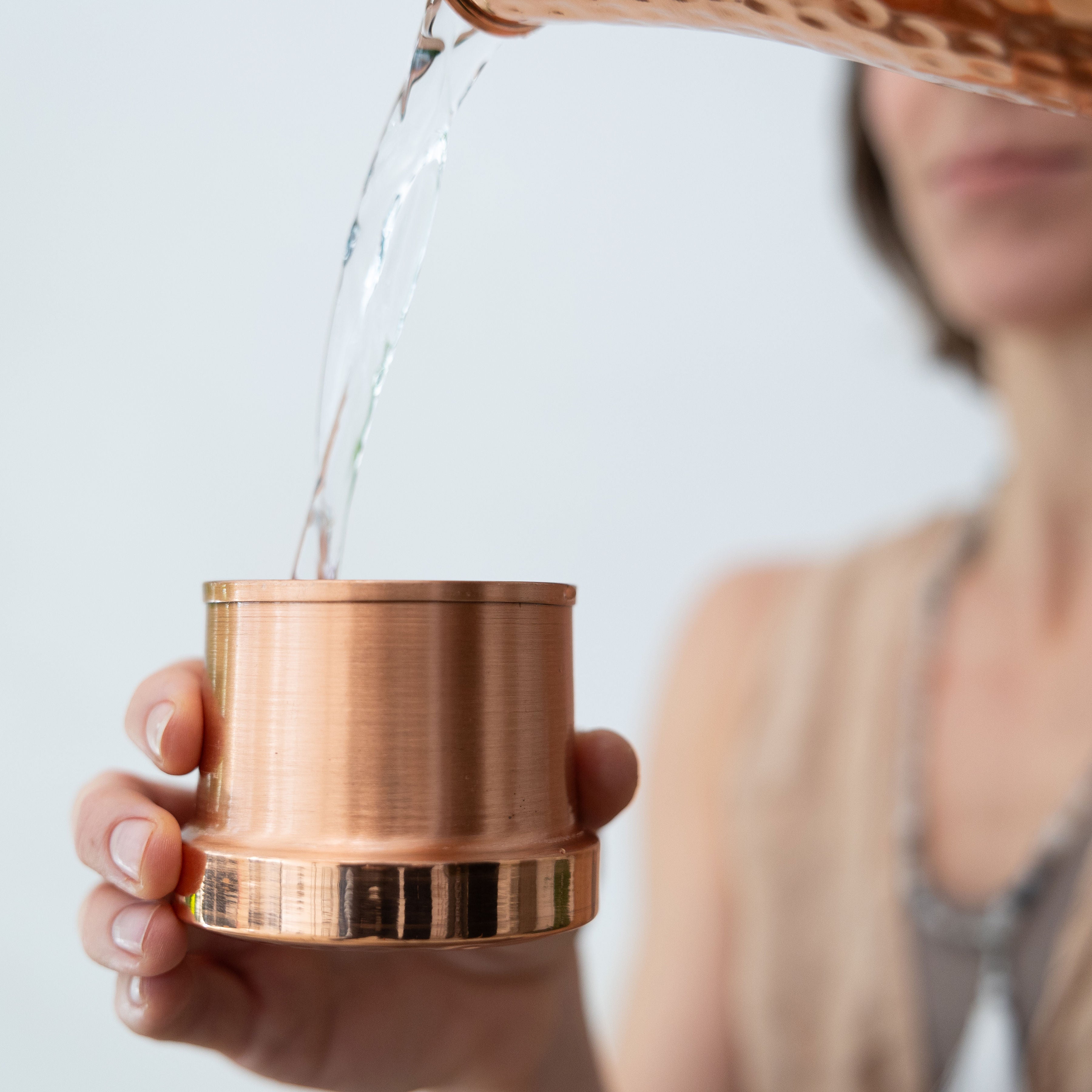 A person pours water from the Maya Shanti Yoga Hammered 950ml Copper Carafe into a pure copper cup with their left hand.