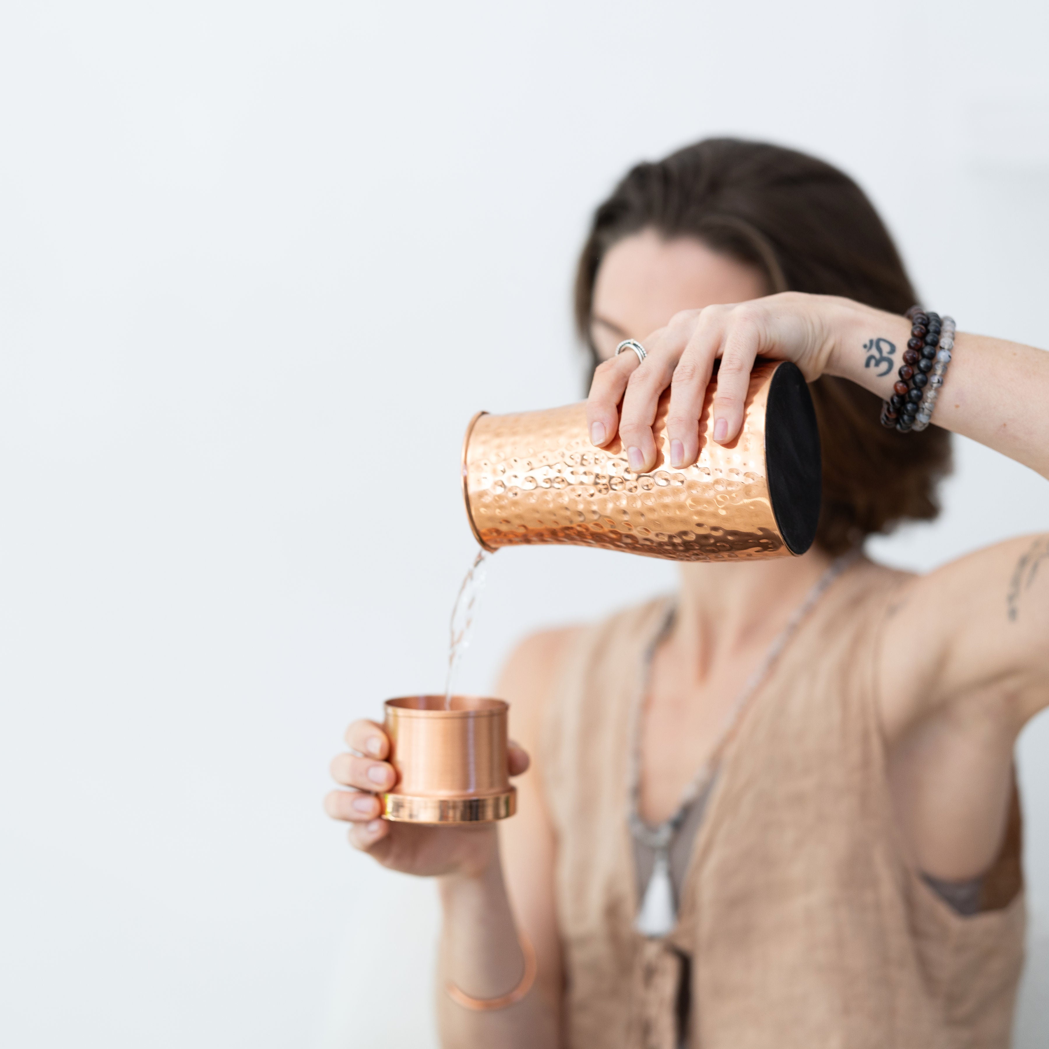 A person pours water from the Maya Shanti Yoga Hammered 950ml Copper Carafe into a small copper cup, wearing Mala Bracelets and Mala Meditation Beads.