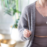 A person wearing a grey sports outfit, long cardigan and Dragon's Eye Mala Meditation Beads holding a lit incense stick in front of them.