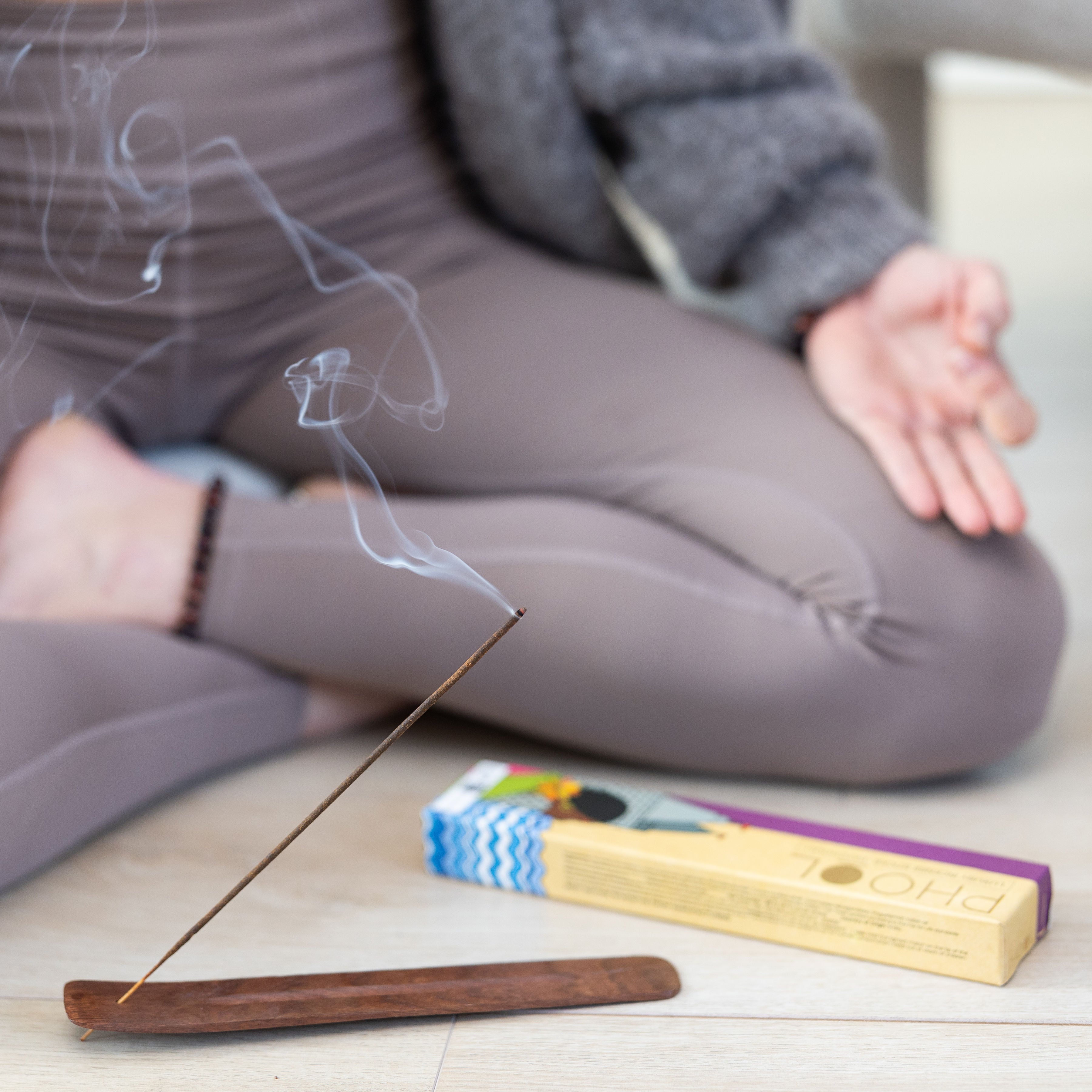 Phool Lavender Incense Sticks burning on a wooden holder with a person in yoga attire meditating cross-legged in the background.