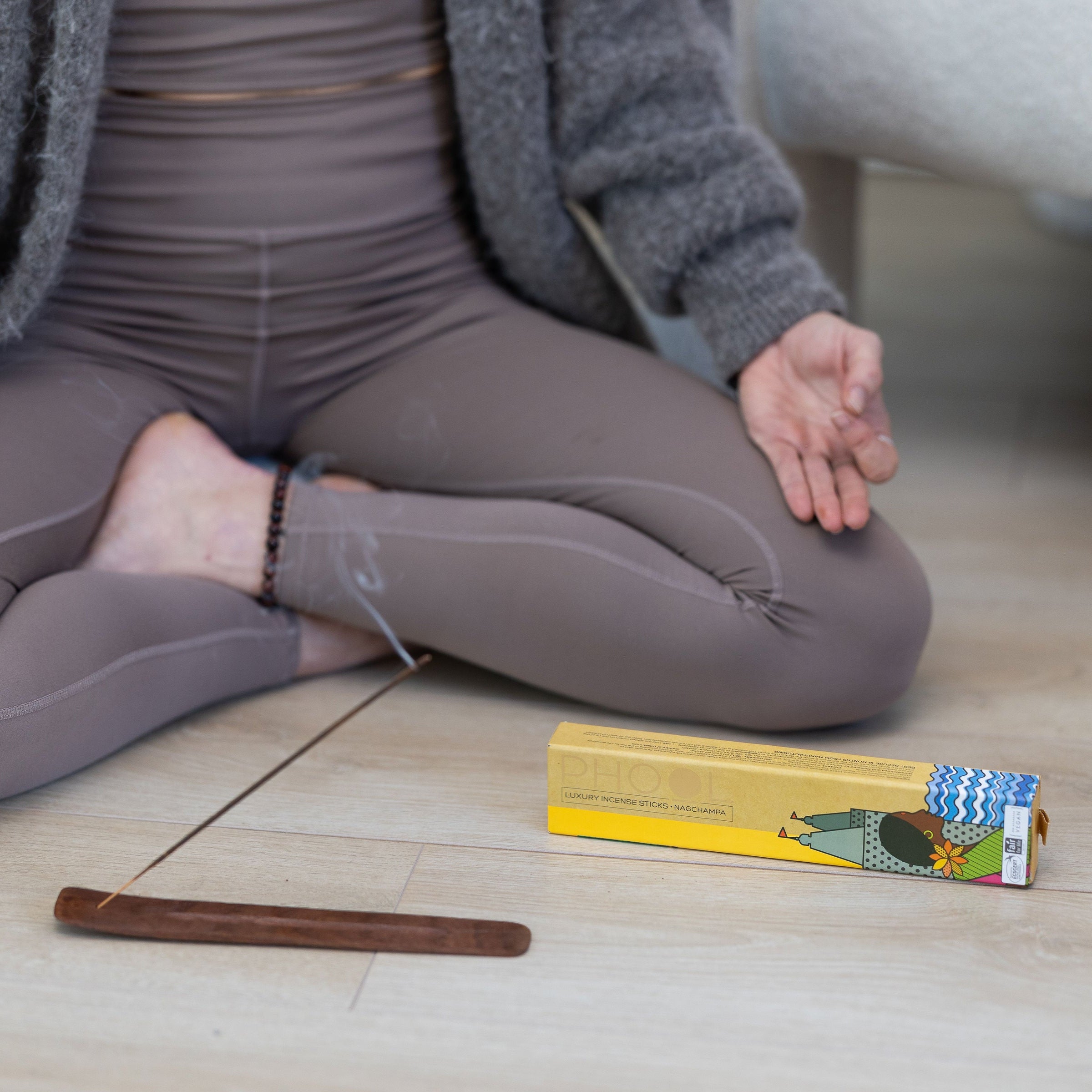 Phool Nagchampa Incense Stick burning on wooden holder next to packaging with woman sitting cross-legged meditating in the background.