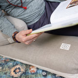 A person reclining comfortably on a Maya Shanti Yoga Zabuton - Meditation Cushion - Hemp & Organic Cotton, engrossed in reading a book.