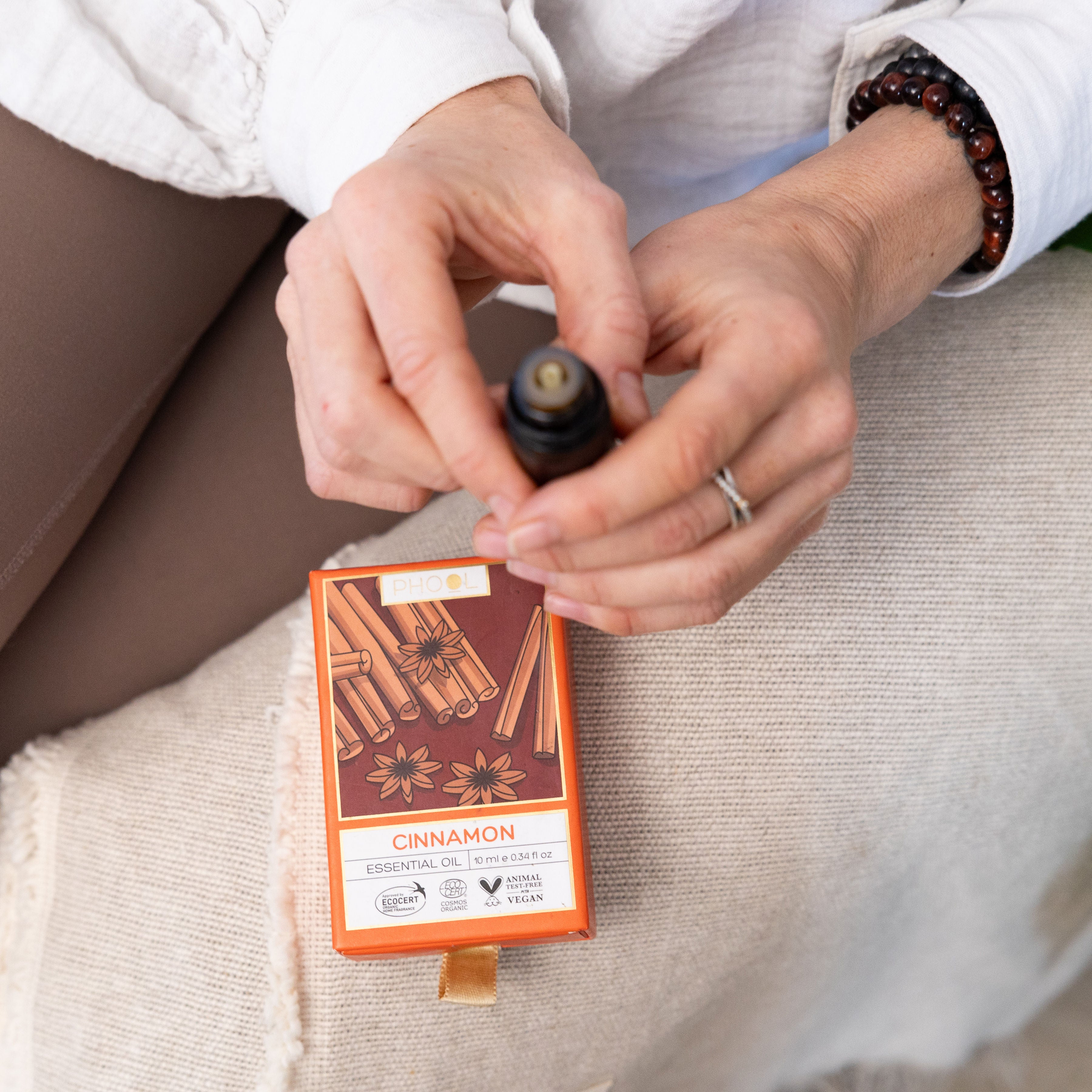 A person holds a small bottle of Phool Cinnamon Essential Oil above an illustrated box while seated on beige fabric.