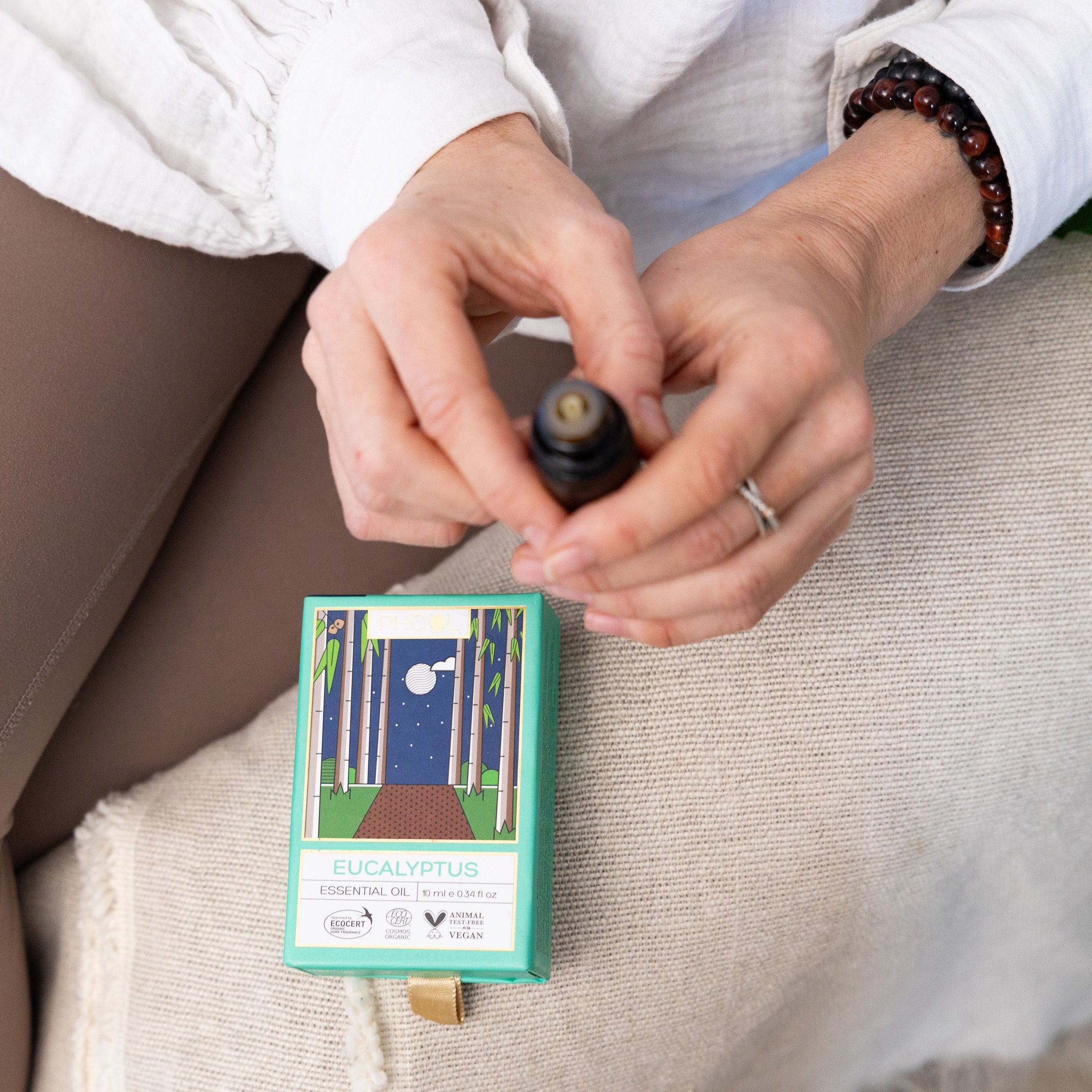 Person holding an open Phool Eucalyptus Essential Oil above the green packaging, while seated on a beige cushion.