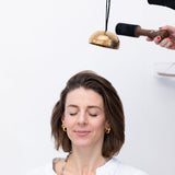 Woman with eyes closed, smiling with another person holding MSY Hanging Himalayan Bowl and wooden mallet above her head.