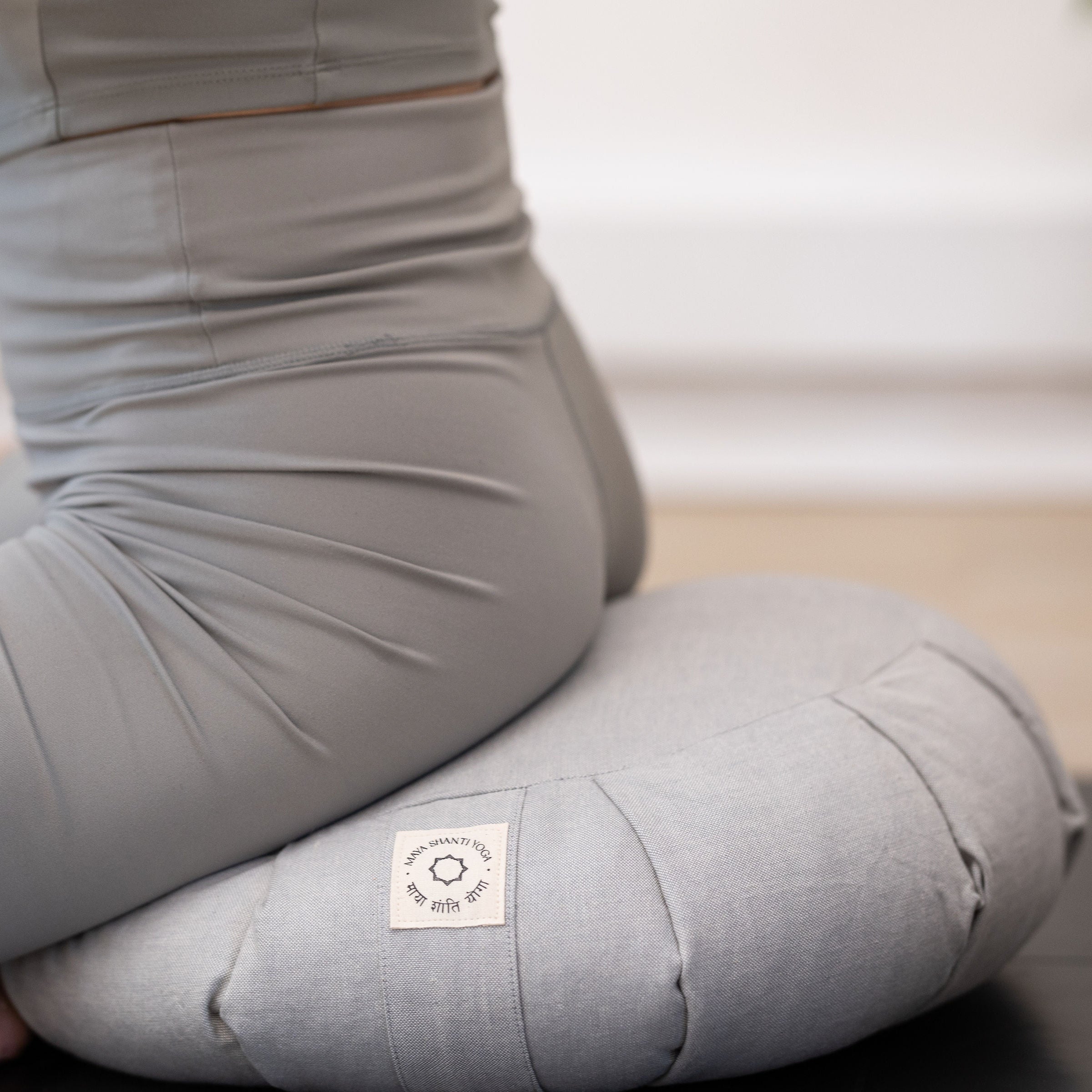 A person in a gray outfit sits cross-legged on an MSY Zafu - Ice Grey by Maya Shanti Yoga, focusing on the lower body. Filled with organic buckwheat hulls, it provides excellent support and stability for yoga meditation.