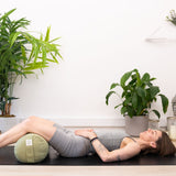 Resting comfortably on a yoga mat, hands on their stomach, a person is surrounded by lush indoor plants and a serene Buddha statue, finding peace with the Maya Shanti Yoga MSY Large Round Buckwheat Bolster in Sage Green for optimal meditation support.