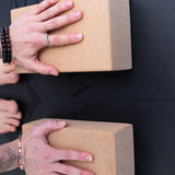 Close-up of 2 hands pressed on cork yoga blocks on a black rubber yoga mat, taken from above.