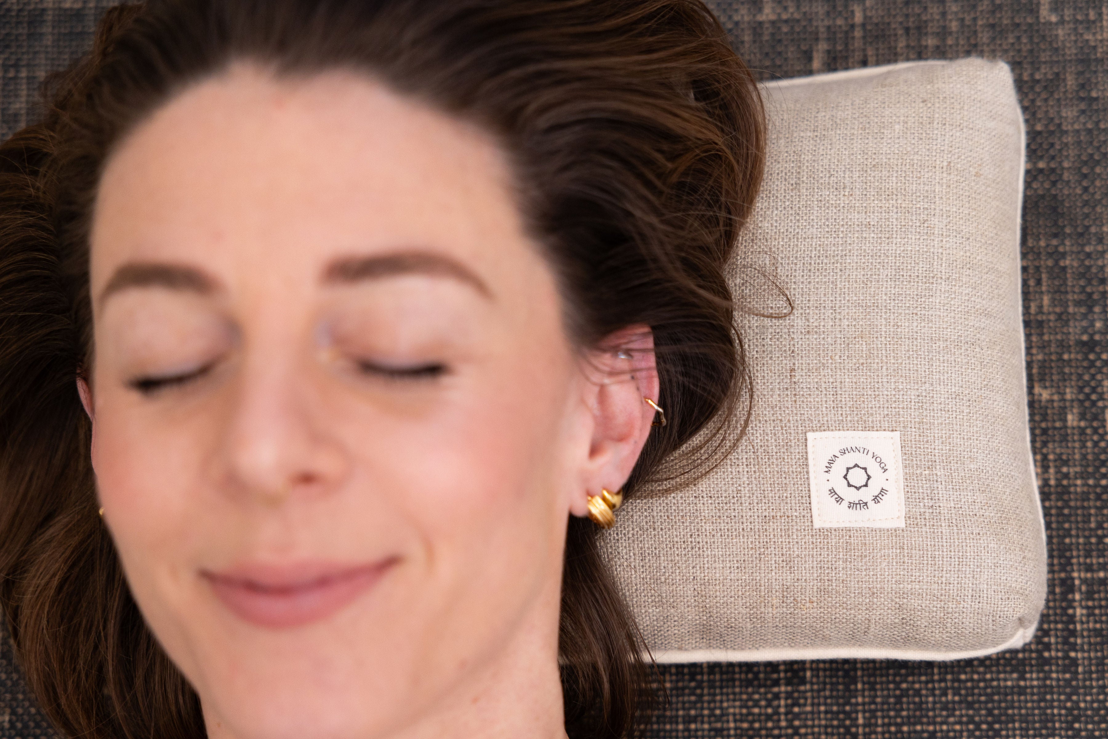 A person with closed eyes rests their head on a beige Maya Shanti Yoga Pillow, made from sustainable hemp with ergonomic support and a small white label, placed on a textured dark surface.