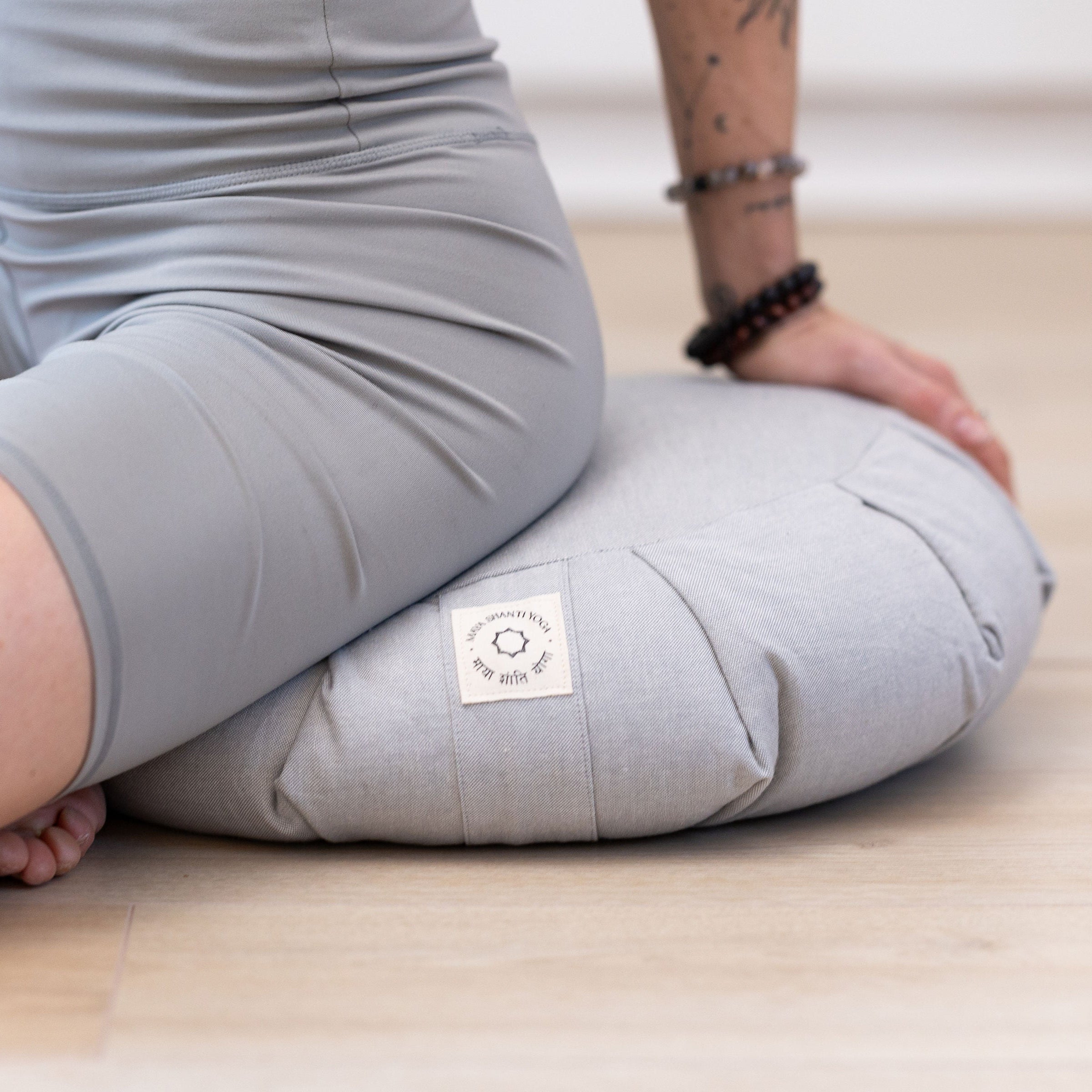 A person in gray workout clothes sits cross-legged on the Maya Shanti Yoga MSY Meditation Zafu – Ice Grey, providing yoga support, atop a light gray cushion on a wooden floor.