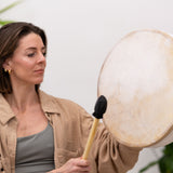 Woman holding MSY Buffalo Drum and drumstick, looking towards it with a soft expression.