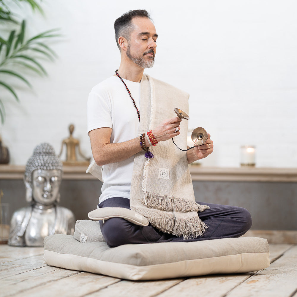 A man sitting cross-legged on cushions in a serene setting holds Tibetan tingsha chimes in his hands. A Buddha statue, a lit candle, and a plant are in the background.