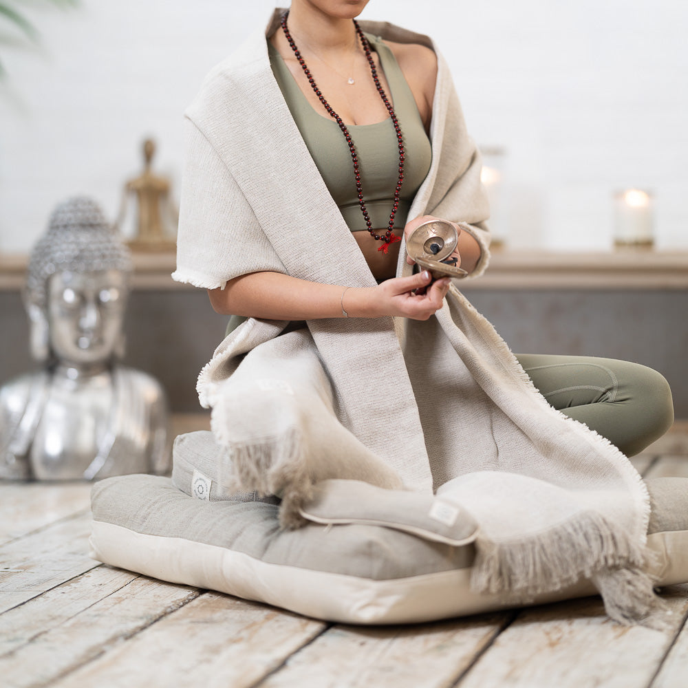 Woman in meditation pose holding a singing bowl, wrapped in a shawl, with a buddha statue in background.