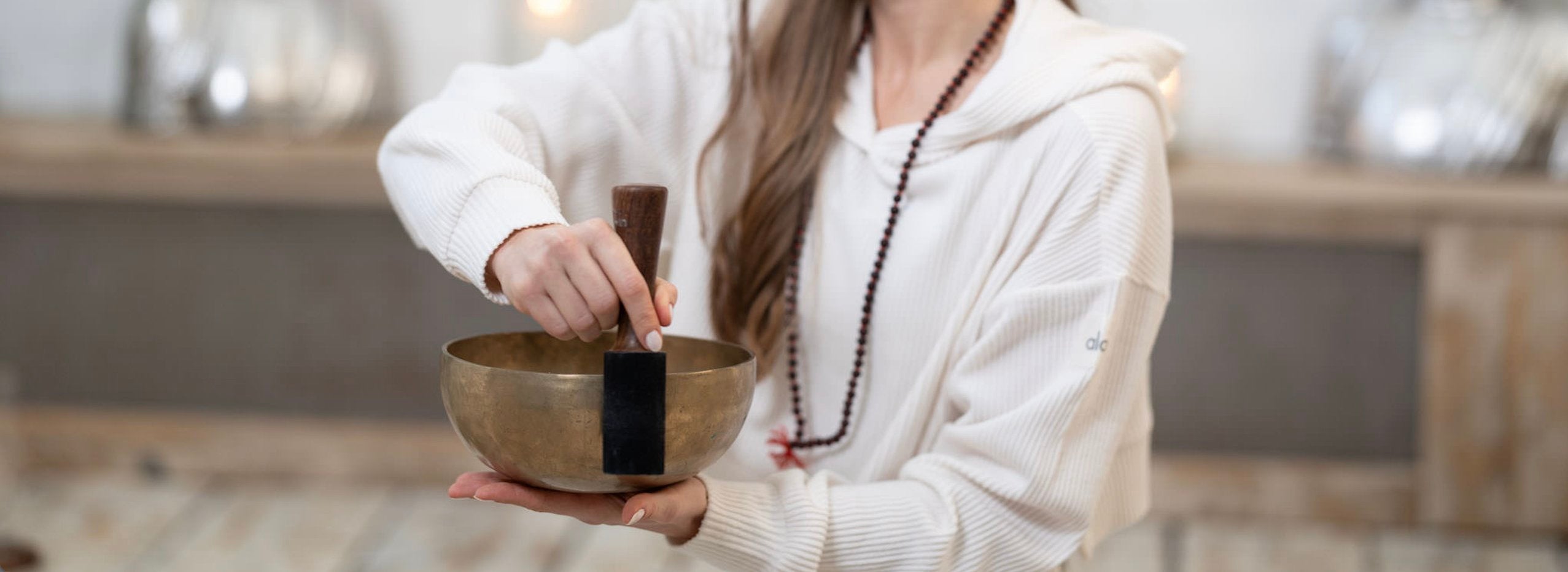 A person wearing a white hoodie holds and rubs a mallet around the rim of a brass singing bowl.