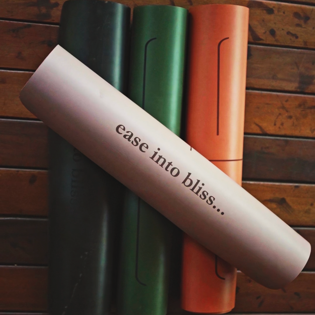 Three MSY Yoga Mats in black, green, and orange on a wooden surface with 'ease into bliss...' text.