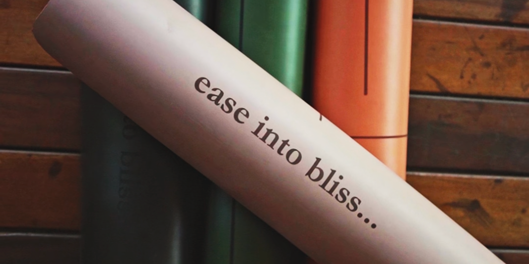 Three MSY Yoga Mats in black, green, and orange on a wooden surface with 'ease into bliss...' text.