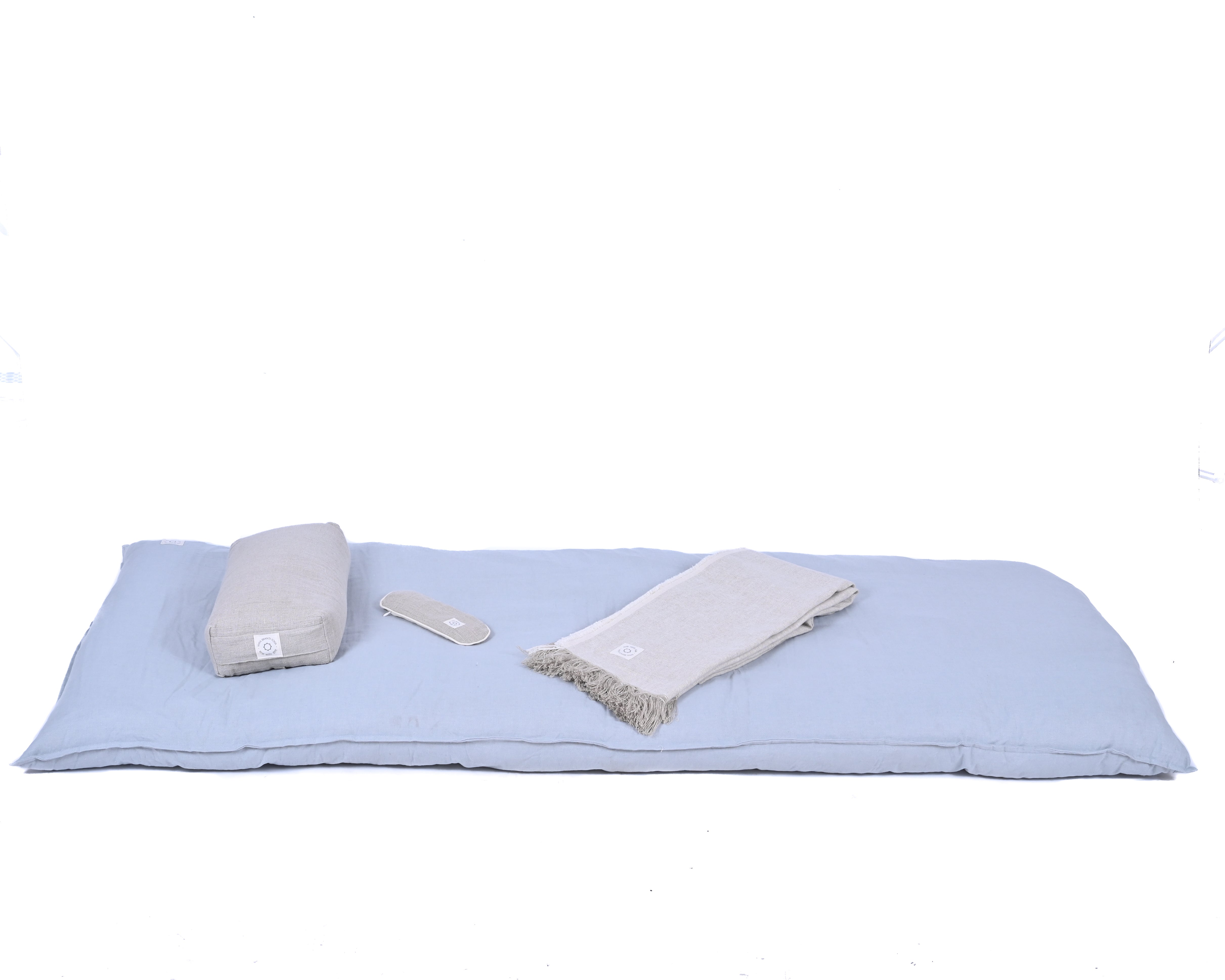 A light blue cushion mat is laid out with a rolled-up gray blanket, a gray eye mask, and a cylindrical gray pillow. There is no detailed product description provided to enhance SEO, but this serene setup promises ultimate comfort and relaxation with the MSY Tranquillity Bundle by Maya Shanti Yoga.