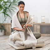 A woman meditating on a cushion, holding a singing bowl, surrounded by candles and MSY Meditation Yoga Blankets in a serene indoor setting. The blankets are made by Maya Shanti Yoga from hemp & organic cotton materials.