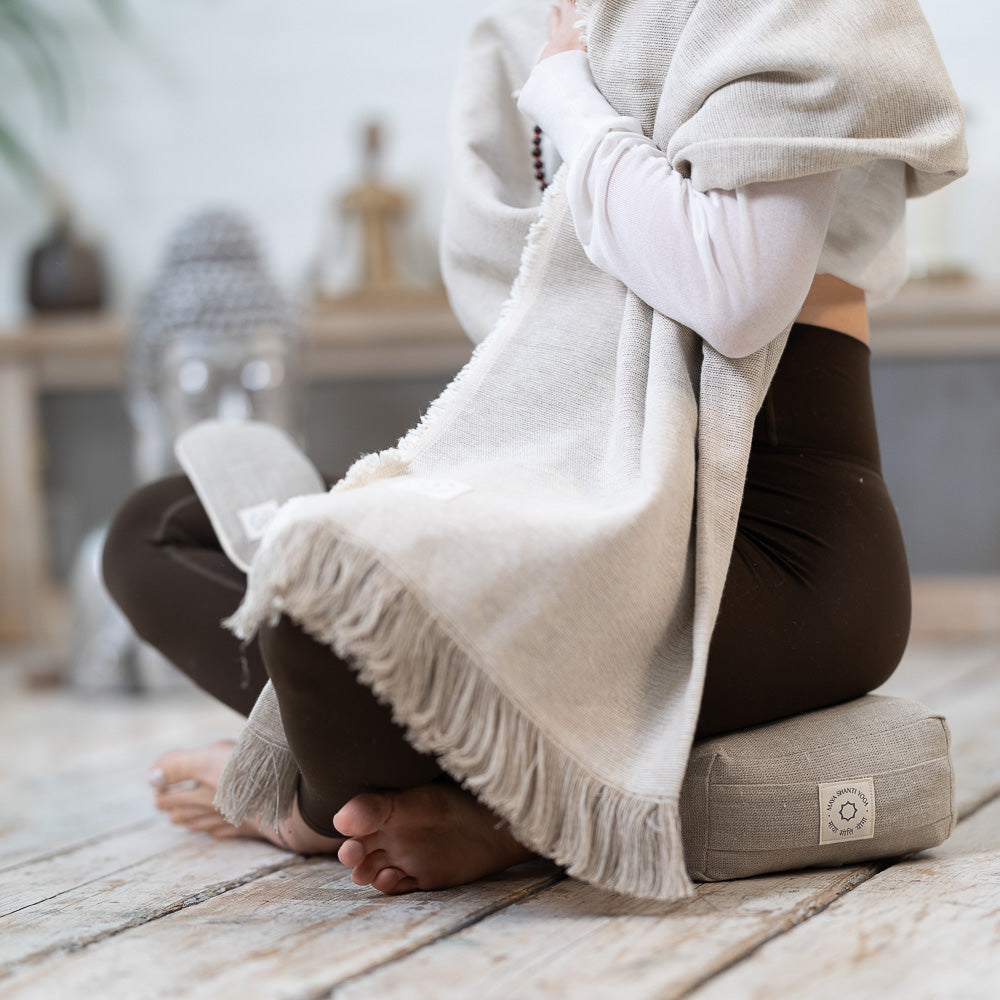 A person sitting cross-legged on a MSY Meditation Yoga Blanket - Hemp & Organic Cotton, draped in a machine-washable shawl, with focus on bare feet and a serene indoor setting. (Brand Name: Maya Shanti Yoga)