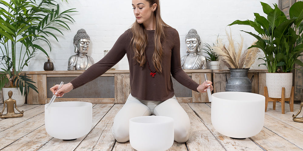 A woman practicing mindfulness and meditation, sitting serenely on a wood floor surrounded by MSY Crystal Singing Bowls- Set of 3 from Maya Shanti Yoga.