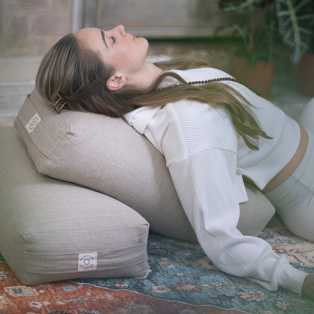 A woman reclining on a Maya Shanti Yoga large rectangular yoga bolster with her head tilted back, eyes closed, wearing a white top and light trousers.
