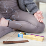 A person in yoga attire meditates cross-legged as a Phool Incense - Lavender by Maya Shanti Yoga burns on a wooden holder, filling the space with its soothing aroma.