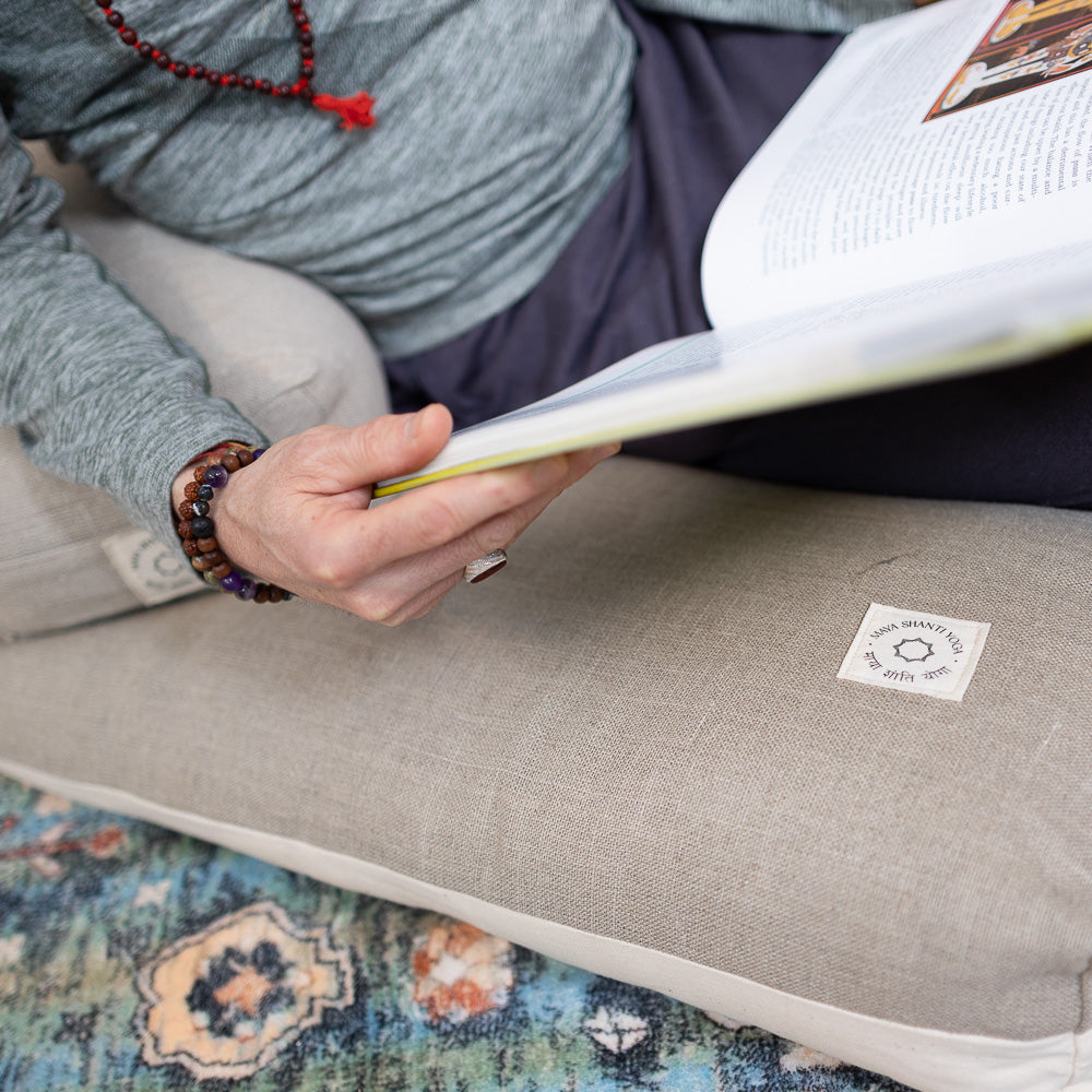 A person reclining comfortably on a Maya Shanti Yoga Zabuton - Meditation Cushion - Hemp & Organic Cotton, engrossed in reading a book.