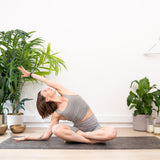 Wearing athletic wear, a person sits indoors on the Maya Shanti Yoga Bengal Yoga Mat Eco-Friendly Extra Grip (Natural Jute and Rubber), performing a seated side stretch. The mat provides a non-slip surface. Potted plants and bowls complete the scene.