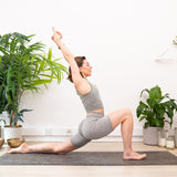 A woman practices yoga indoors, performing a low lunge with arms raised on the Maya Shanti Yoga Bengal Yoga Mat Eco-Friendly Extra Grip, made from natural jute and rubber, surrounded by potted plants.