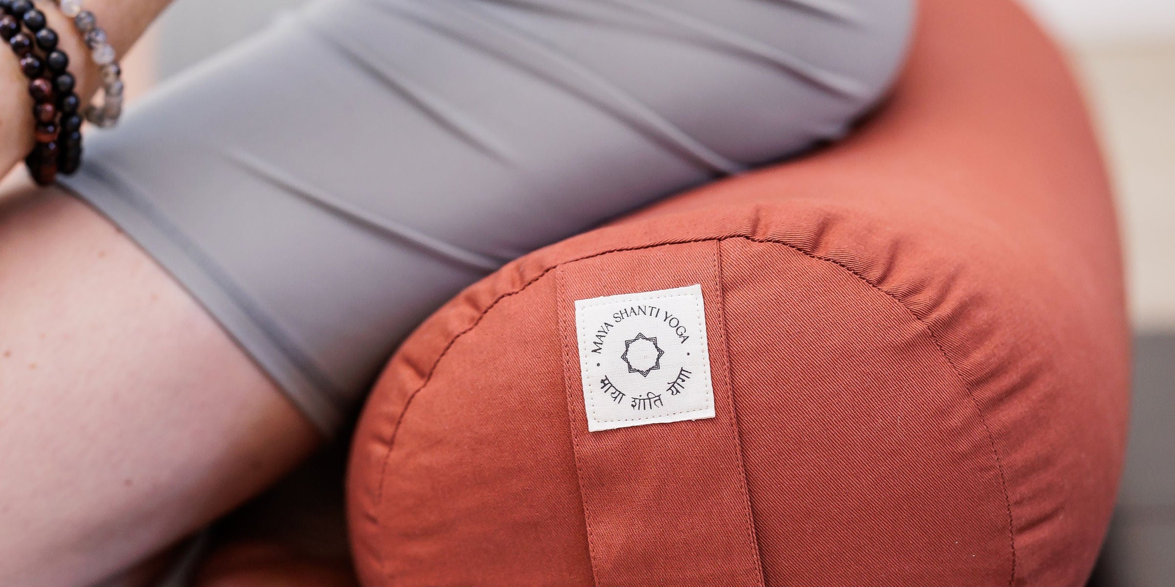 A person wearing gray sits on a Maya Shanti Yoga MSY Large Round Buckwheat Bolster in Rust, hands on knees revealing a tattooed arm.