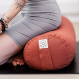 A person wearing gray sits on a Maya Shanti Yoga MSY Large Round Buckwheat Bolster in Rust, hands on knees revealing a tattooed arm.