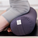 A person in gray athletic wear kneels on a black mat, practicing yoga with the eco-friendly Maya Shanti Yoga MSY Large Round Buckwheat Bolster in Anthracite Grey.