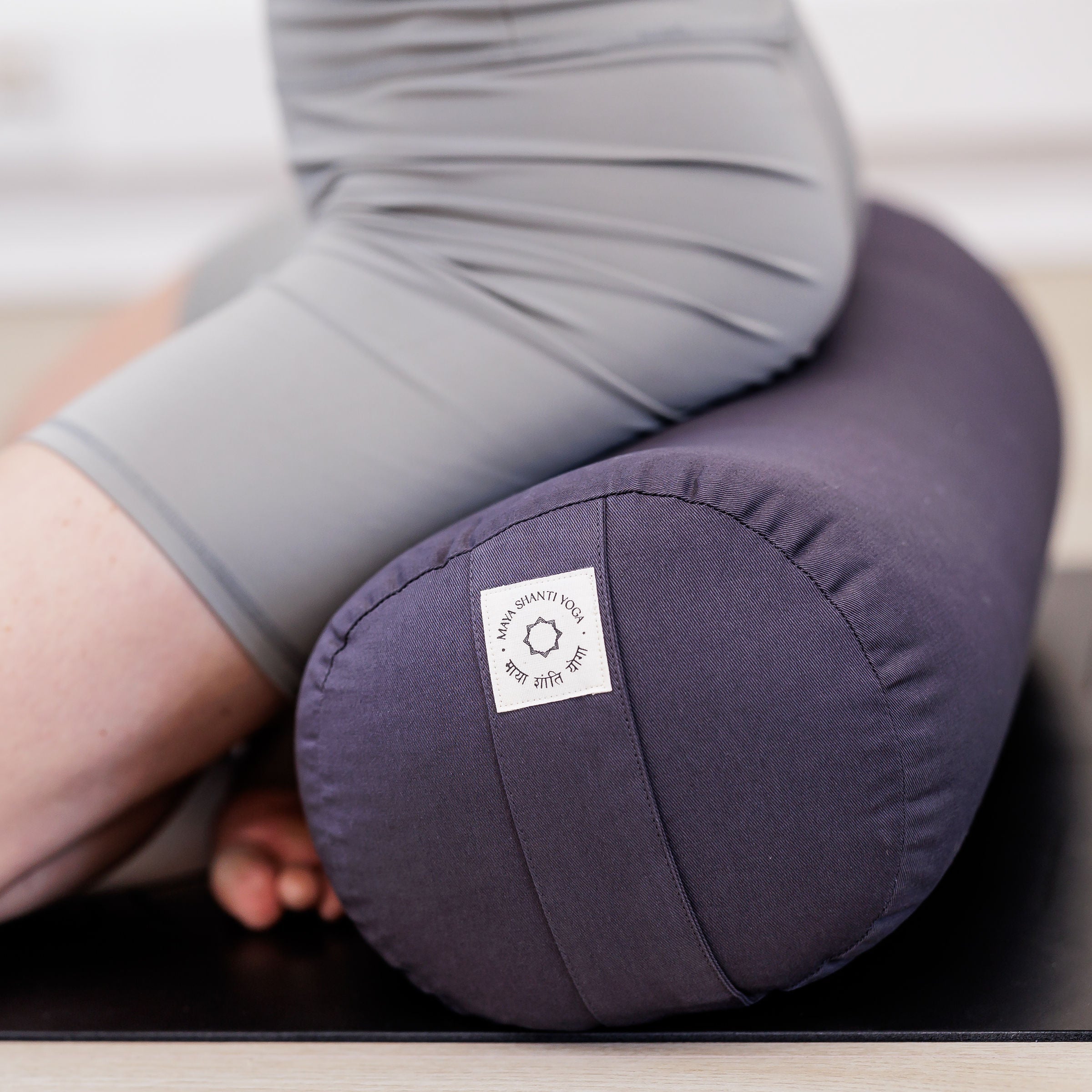 A person in gray athletic wear kneels on a black mat, practicing yoga with the eco-friendly Maya Shanti Yoga MSY Large Round Buckwheat Bolster in Anthracite Grey.