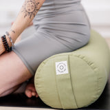 A calm person in a gray outfit kneels on their yoga mat, adorned with beaded bracelets and rings, showcasing a sun tattoo. In front of them lies the MSY Large Round Buckwheat Bolster in Sage Green by Maya Shanti Yoga, providing natural support to their serene practice.