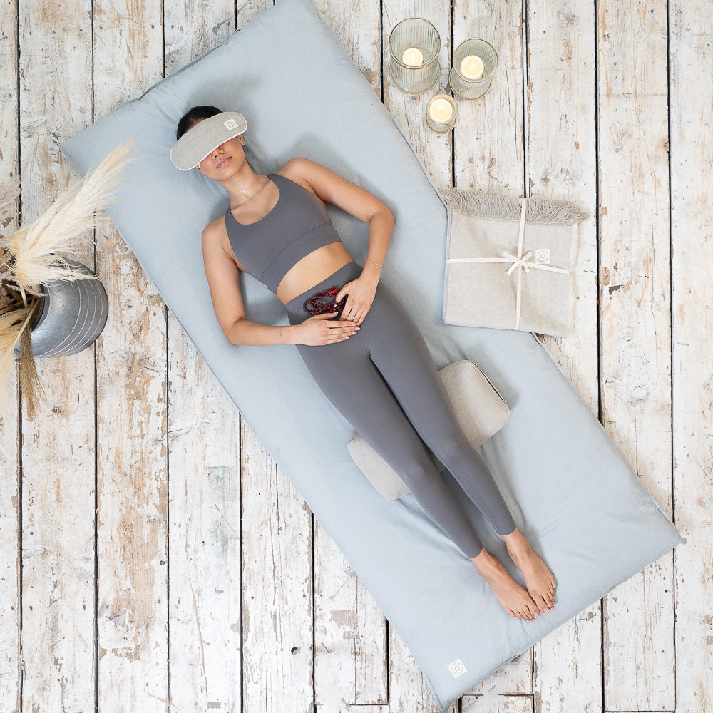 A woman practicing mindfulness and wellness, lying on a MSY Shiatsu Meditation Bed - Organic Cotton from Maya Shanti Yoga and wearing an eye mask for relaxation.