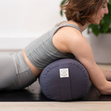 A person in a gray workout outfit is lying on a yoga mat, using the Maya Shanti Yoga MSY Large Round Buckwheat Bolster - Anthracite Grey, filled with organic buckwheat hulls, under their torso.