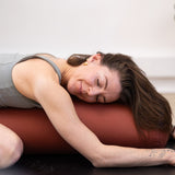 A person reclines on a Maya Shanti Yoga MSY Large Round Buckwheat Bolster - Rust, eyes closed in a relaxed pose, wearing a gray athletic outfit.