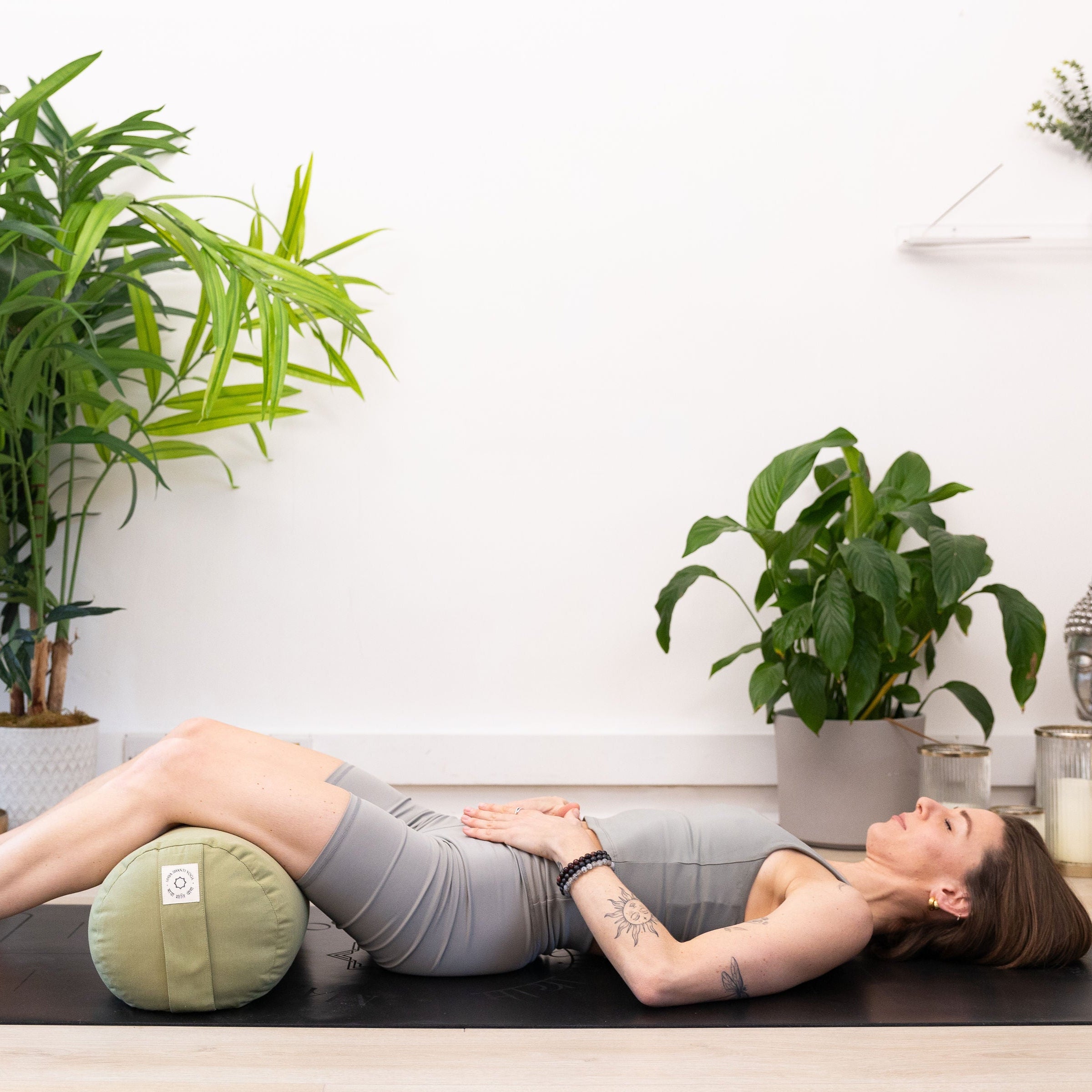 Resting comfortably on a yoga mat, hands on their stomach, a person is surrounded by lush indoor plants and a serene Buddha statue, finding peace with the Maya Shanti Yoga MSY Large Round Buckwheat Bolster in Sage Green for optimal meditation support.