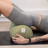 A person in gray athletic wear performs a bridge pose on a yoga mat, supported by the eco-friendly Maya Shanti Yoga MSY Large Round Buckwheat Bolster in Sage Green, with tattoos visible on their arm, enhancing the artistry of their practice.