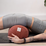 A person in gray athletic wear lies on a yoga mat, arching over a MSY Large Round Buckwheat Bolster - Rust by Maya Shanti Yoga, filled with organic buckwheat hulls, arms wrapped around it. A tattooed arm is visible.